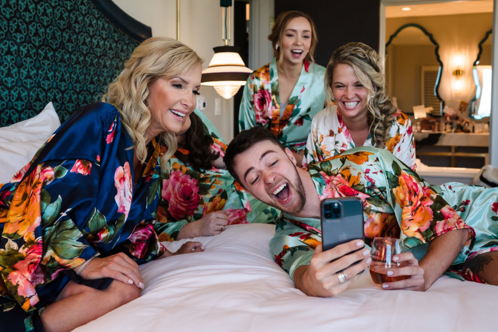 Bride, bridesmaids, bridesman, and bride's mother laughing at phone in hotel room in Uptown Charlotte