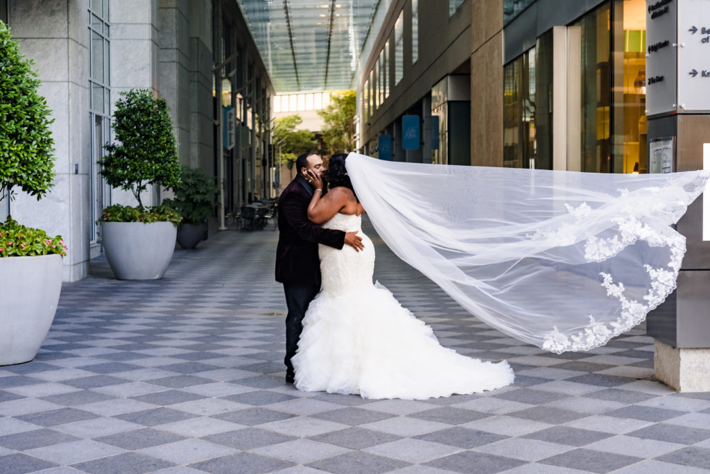 Veil blowing in the wind while a bride and groom kiss in front of an alley in Uptown Charlotte right next to the Mint Museum.