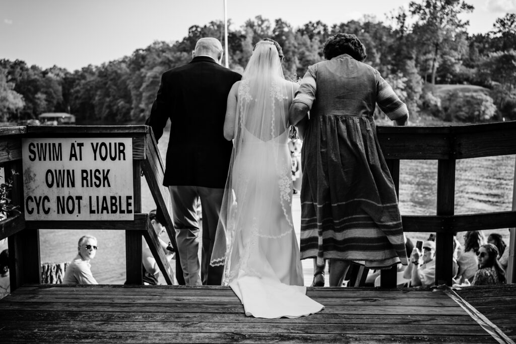Black and White photo of the bride being walked to a lakefront intimate wedding ceremony