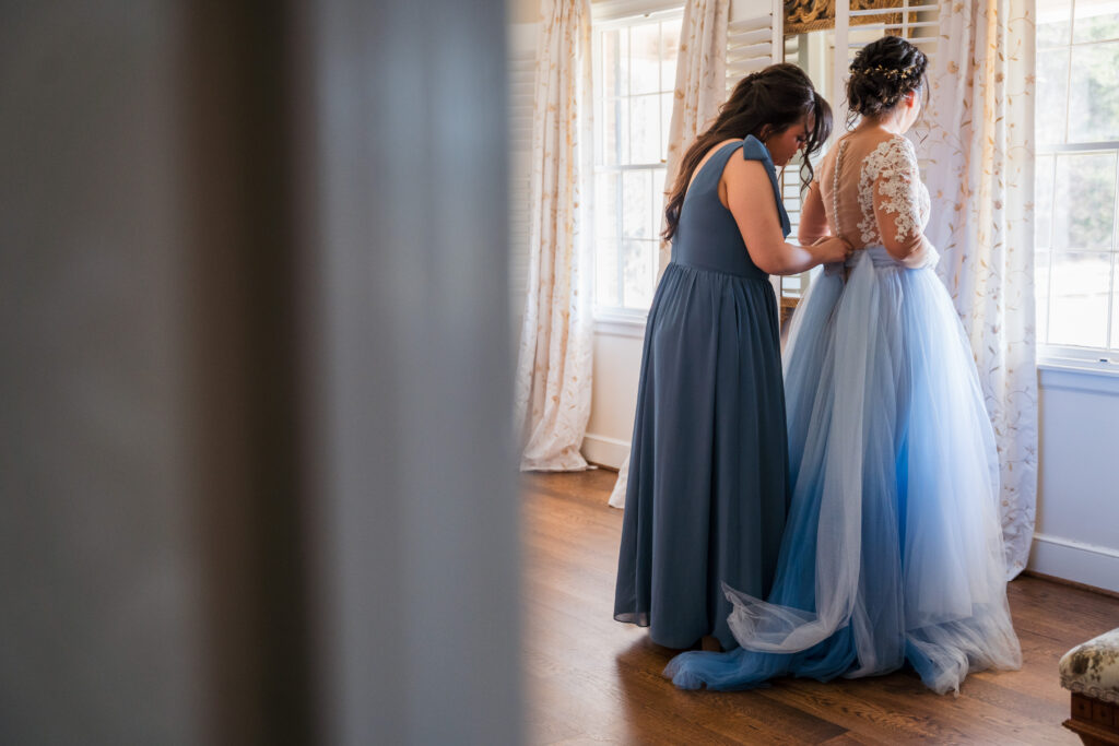 A bridesmaid wearing a blue dress helping the bride get her dress on upstairs inside the home on Pleasant Grove Farm's property