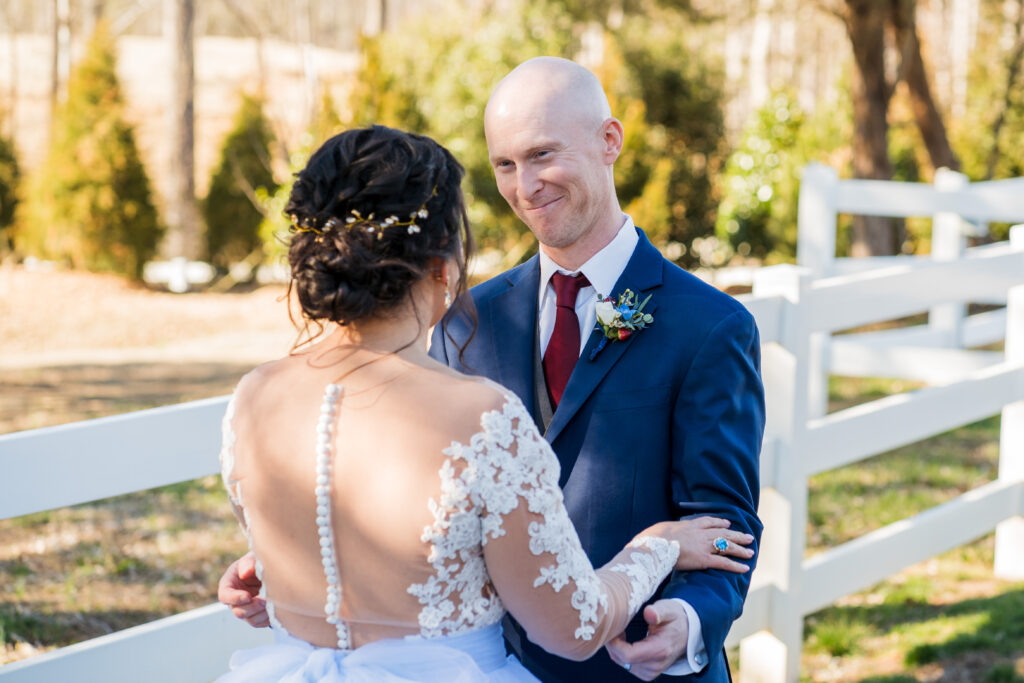 Groom smiles at bride after first look outside at Pleasant Grove Farm in front of white picket fence