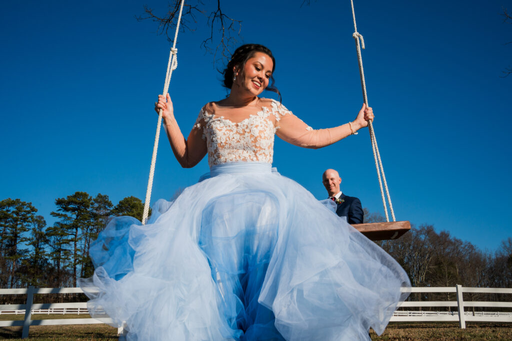 Groom pushes bride on tree swing outside at Pleasant Grove Farm with blue sky and blue dress