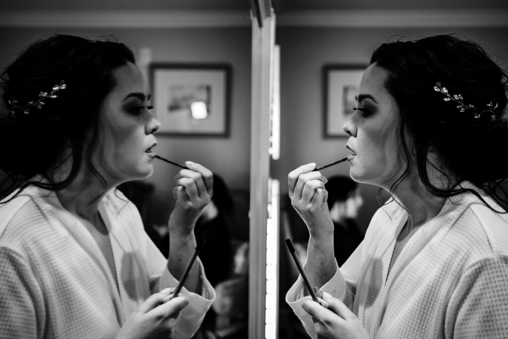 A bride looking in the mirror as she puts her lip liner on, in black and white