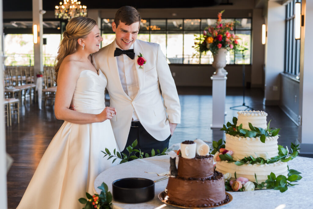 Bride and Groom dressed in white see the cake with colorful Spring flowers before their wedding at Terrace at Cedar Hill