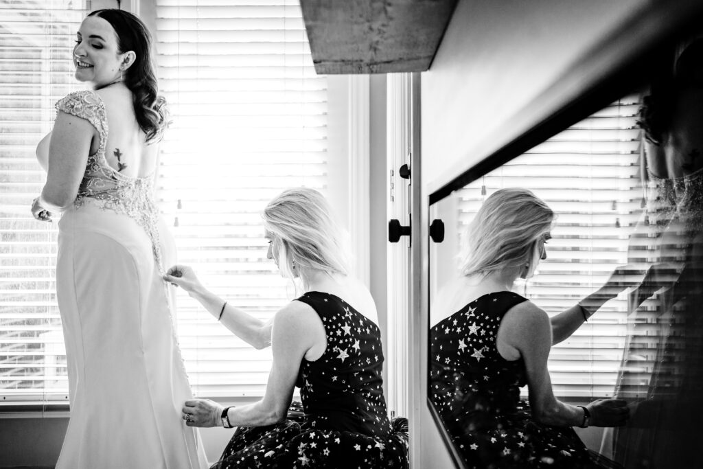 Black and white wedding photo of bridesmaid helping bride get dressed with fireplace reflection